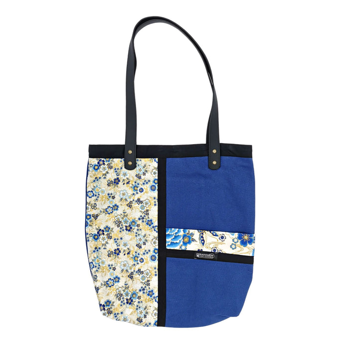 Tote Bag hecha de algodón de color azul. Tiene una estampa que cubre el lado derecho con un diseño japonés de flores. Las tonalidades son azules, blancas y amarillas. Posee dos asas de cuerina negra de 70 cm.