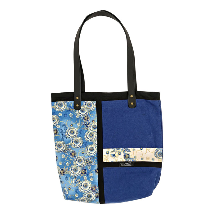 Tote Bag hecha de algodón de color azul. Tiene una estampa que cubre el lado derecho con un diseño de flores y líneas de color amarillo, blanco y tonos azules. Posee dos asas de cuerina negra de 70 cm.