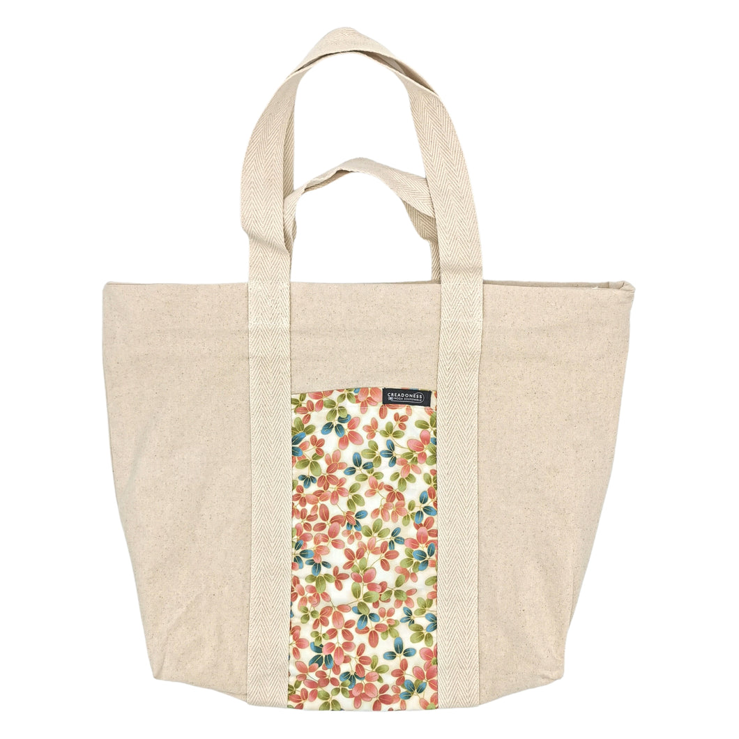 Maxi Bag hecha de algodón de color blanco con un bolsillo exterior de color beige y diseño floral. Las flores son pequeñas y de color durazno, azul y verde. Su medida es de 46 cm de alto